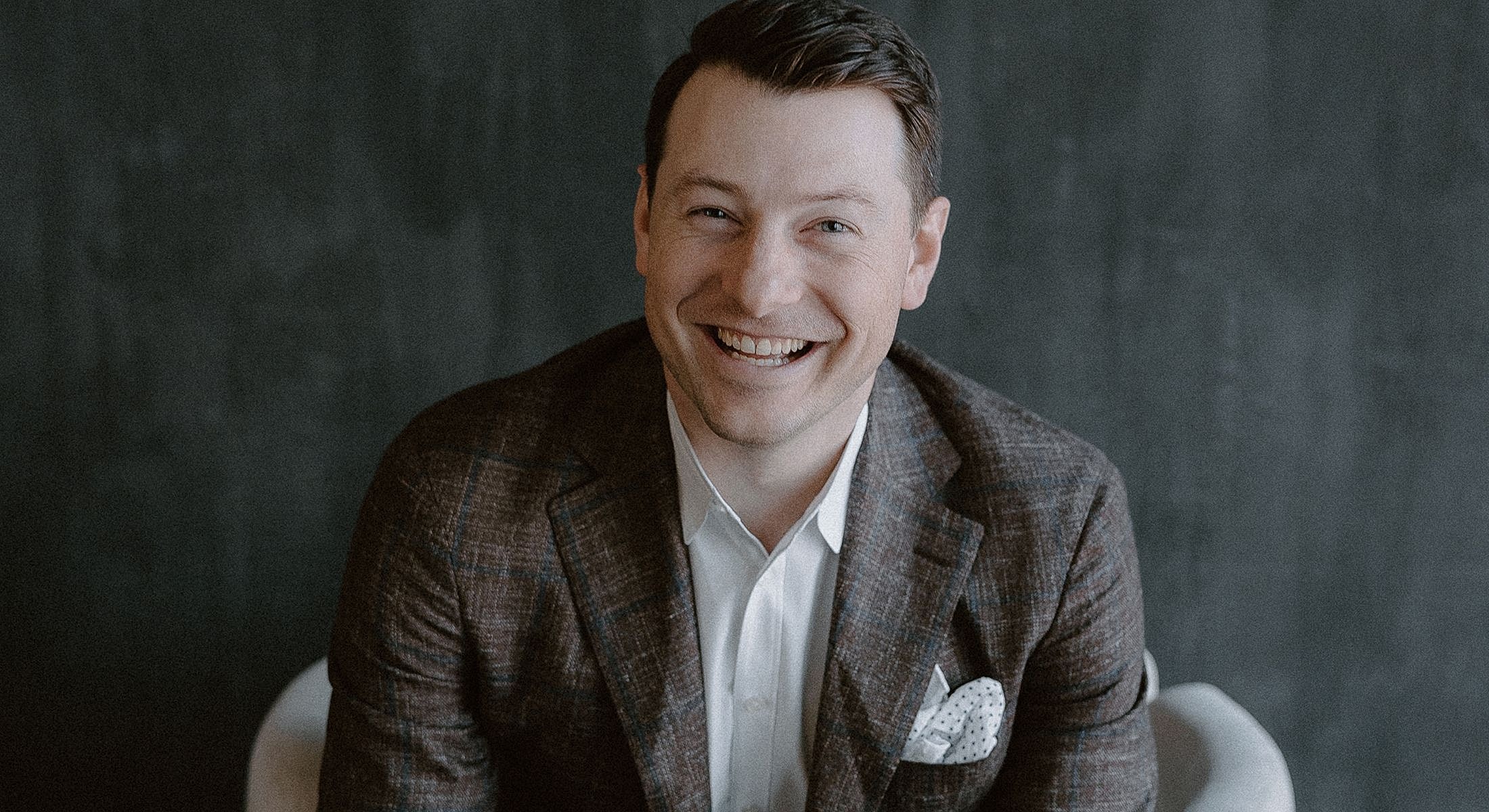 Smiling man in a blazer against a dark background.